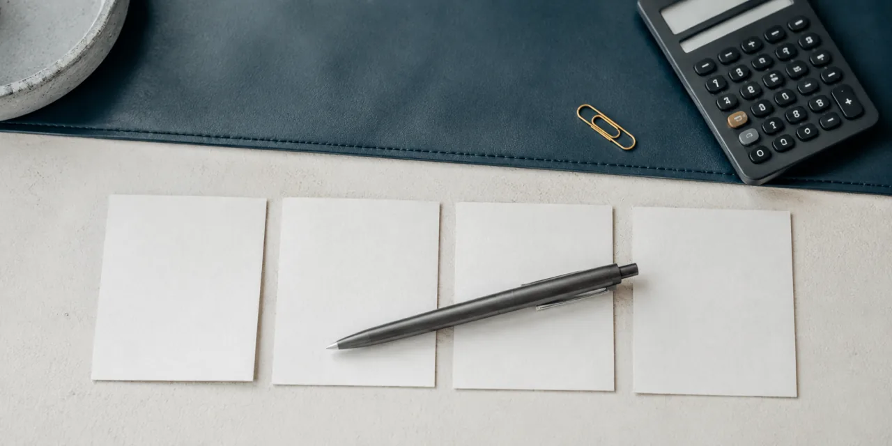 Quatre petites cartes de papier vierges alignées sur un bureau, un stylo posé en travers, une calculatrice et un trombone ocre au bord d’un sous-main bleu ardoise